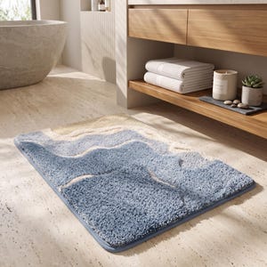 May include: A rectangular bath mat featuring a textured, shaggy pile surface with a wavy, abstract pattern in shades of blue, cream, and white. The mat has a finished edge border and is designed for placement on a bathroom floor surface.