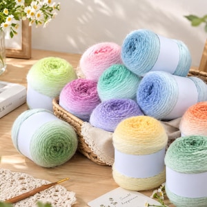 Könnte beinhalten: Eine Sammlung weicher, flauschiger Garnknäuel in verschiedenen Farbverläufen wie Pink, Lila, Blau, Gelb und Grün, jeweils mit einer schlichten weißen Papierbanderole versehen, angeordnet in einem Weidenkorb auf einer Holzoberfläche.