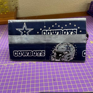 May include: A rectangular fabric clutch featuring a navy blue and grey color scheme with the Dallas Cowboys logo, a football helmet graphic, and the text COWBOYS. The design is accented with small clear rhinestones across the surface.