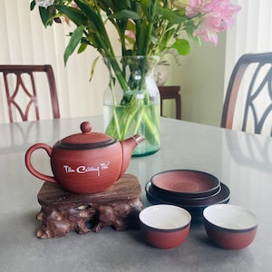 Peut inclure: Un service à thé en terre cuite comprenant une théière ronde avec couvercle portant l'inscription blanche "Tân Cương Trà", deux petites tasses assorties à l'intérieur blanc et deux soucoupes marron foncé, posé sur un socle en bois.