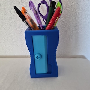 Peut inclure: Un pot à crayons bleu foncé en forme de taille-crayon avec un panneau central bleu clair doté d'une petite vis en forme de croix. Le pot contient diverses fournitures de bureau, dont un stylo noir, un surligneur orange, un stylo violet, des ciseaux violets, un stylo vert foncé et un stylo rouge.