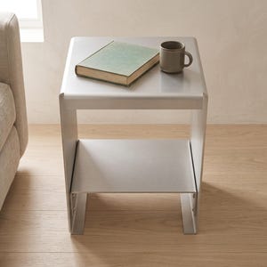 May include: A silver-toned metal side table with a square top and a lower shelf. A light green hardcover book and a small, solid-colored gray ceramic mug are placed on the top surface of the table, which features a smooth, matte finish.