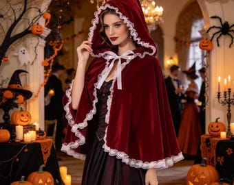 May include: A deep red velvet hooded cape featuring a white lace trim along the edges and a white ribbon tie at the neck. The cape is worn over a dark dress, with the hood pulled up to frame the face, creating a classic, layered appearance.