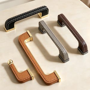 以下が含まれることがあります： 白いコントラストステッチが施されたレザー調のテクスチャー素材を使用したキャビネットプルとノブのコレクション。金具はブラック、ブラウン、グレー、タンのカラー展開で、ゴールドまたはダークグレーの金属製エンドキャップと取り付けベースが付いています。