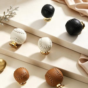 May include: A collection of six spherical cabinet knobs shaped like baseballs, featuring textured leather surfaces in black, white, and brown. Each knob is attached to a polished gold-tone metal base, showcasing detailed stitching patterns.