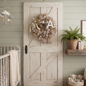 May include: A decorative wreath made of tan burlap mesh, featuring a plush tan bunny, white star accents, a large white letter A, and a tan and white checkered ribbon bow, mounted on a white distressed wooden barn door with a black metal handle.