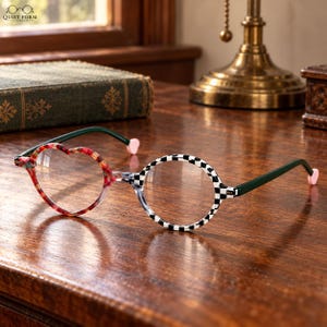 May include: Asymmetrical eyeglasses featuring one heart-shaped lens frame with a red and brown tortoiseshell pattern and one circular lens frame with a black and white checkerboard pattern. The temples are dark green with small pink heart-shaped tips.