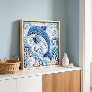 Könnte beinhalten: Ein quadratisches Wandbild aus Stoff in einem hellen Holzrahmen mit einem gesteppten Delfin in Blau- und Weißtönen. Das Design umfasst wirbelnde Wellenmuster, Korallenstrukturen und Muscheln, die alle aus verschiedenen blauen Stoffen gefertigt sind.