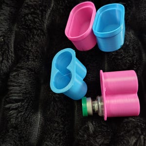 May include: A collection of four plastic holders in bright pink and blue colors. Two are single oval-shaped cups, one is a double-lobed blue holder, and one is a double-lobed pink holder containing a small glass vial with a green cap.