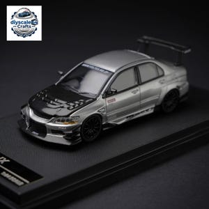 May include: A silver 1:64 scale model car featuring a black carbon fiber hood, a rear spoiler, and black multi-spoke wheels. The vehicle displays various sponsor decals, including PIAA and Motul, with the text WWW.VOLTEX.NE.JP on the windshield.
