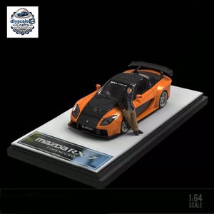 Może przedstawiać: Model samochodu sportowego Mazda RX-7 w skali 1:64 w kolorze pomarańczowo-czarnym z zestawem nadwozia VeilSide. Mała figurka opiera się o maskę. Samochód jest zamontowany na podstawie z napisem Mazda RX-7 TIMEMICRO oraz numerem seryjnym 001-999.