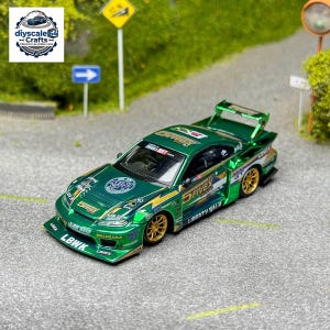 May include: A metallic green die-cast model car featuring gold wheels, a rear spoiler, and various sponsor decals including Fivex, Liberty Walk, and Mini GT. The car has a low-profile body kit and is displayed on a miniature asphalt road surface.