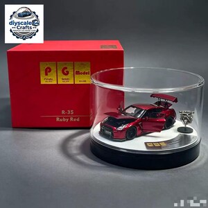 May include: A ruby red R-35 model car with open doors and bonnet, displayed inside a clear cylindrical case on a black base. Next to the car is a small silver engine component. The set includes a red box labelled R-35 Ruby Red and PG Model.
