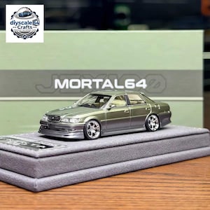 Può includere: Un modellino di auto in scala 1:64 con finitura bicolore verde oliva metallizzato e argento. La berlina presenta cerchi cromati a più razze, fari trasparenti e un paraurti anteriore ribassato su una base rettangolare grigia.
