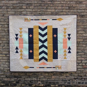 May include: A geometric quilt with a white background and a pattern of black, gold, and light blue triangles, squares, and chevron shapes. The quilt also features peach and light blue rectangular shapes.