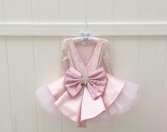 May include: A pink tulle and satin dress with a large pink satin bow and a jeweled embellishment. The dress has long sleeves and a lace bodice.