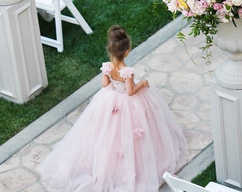 May include: A young girl in a light pink tulle dress with floral accents walks along a stone path. The dress features a full skirt and delicate details. The scene is set outdoors with greenery and white chairs in the background.