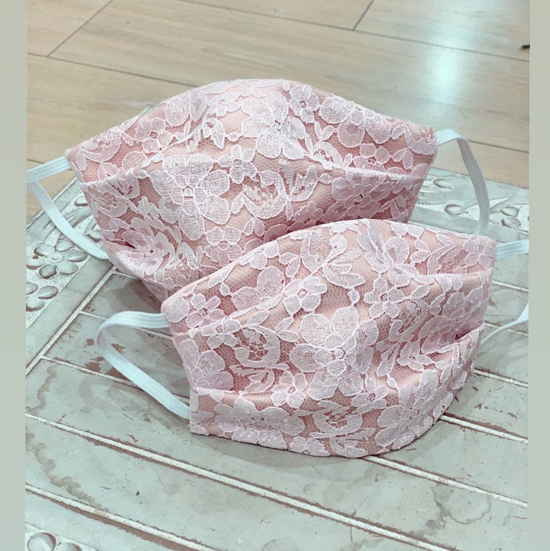 May include: Two pink lace face masks with white elastic straps. The masks are made of delicate lace fabric with a floral pattern.