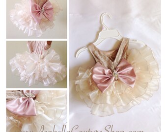 May include: A beige and pink baby girl dress with a large pink satin bow and a rhinestone embellishment. The dress has multiple layers of tulle ruffles.