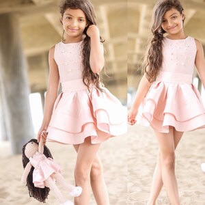 May include: Two young girls wearing pink sleeveless dresses with pearl embellishments and ruffled skirts. The girl on the left is holding a pink doll with brown hair.