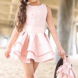 May include: A young girl in a pink dress with pearl embellishments holds a crocheted doll with a pink dress and brown hair. The girl is standing on a sandy beach.