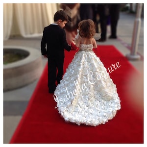 May include: A young girl in a white, ruffled dress walks down a red carpet with a young boy in a black suit. The girl's dress is made of many layers of fabric that resemble roses. The text "Isabella Couture" is visible on the dress.