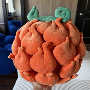 May include: A round, plush toy shaped like a fruit with a vibrant orange exterior covered in flame-like, swirling patterns. The top of the object features a distinct, curved green stem made of soft fabric, adding a contrasting color element.