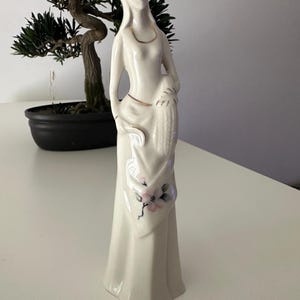 以下が含まれることがあります： 長く流れるようなガウンをまとった立ち姿の、背が高くスリムな白い磁器製の置物。ガウンには、緑の葉を添えた繊細なピンクの花が描かれており、ネックラインとウエストには細い金の縁取りが施されています。