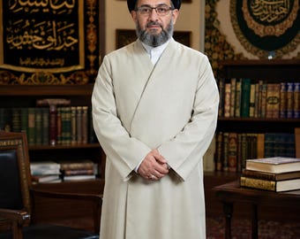May include: A person wearing a light beige, long-sleeved, floor-length robe with a high collar and a black turban. The person has a full beard, wears glasses, and has a silver ring on their finger. The attire is simple, solid-colored, and formal.