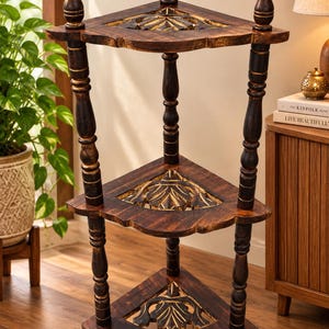 May include: A three-tier corner shelf crafted from dark brown wood with a distressed finish. Each triangular shelf features a carved leaf motif in the centre. The unit is supported by four turned wooden pillars with decorative finials at the top.