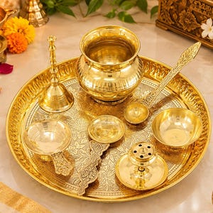 May include: A polished brass puja thali set featuring a large circular tray with intricate floral engravings, a central kalash pot, a bell, a spoon, two small bowls, and a diya lamp, all arranged on a reflective metallic surface.