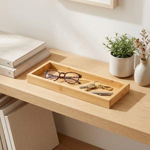 Puede incluir: Una bandeja rectangular de bambú con acabado de veta de madera clara. Dentro de la bandeja hay un par de gafas con patrón de carey marrón, un llavero de metal con varias llaves y un bolígrafo de metal plateado colocado horizontalmente.