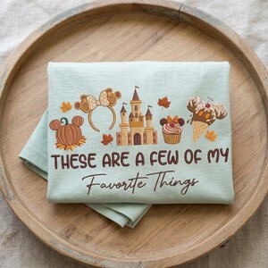 May include: A light green t-shirt featuring embroidered icons of a pumpkin, mouse ears headband, castle, cupcake, and ice cream cone, with the text "THESE ARE A FEW OF MY Favorite Things" in brown lettering and scattered autumn leaves.