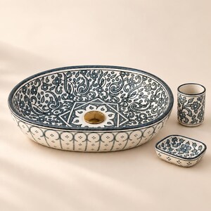 Könnte beinhalten: Ein ovales Aufsatzwaschbecken aus Keramik mit blau-weißem Blumenmuster, einem zentralen geometrischen Motiv und einem goldfarbenen Abfluss. Dazu gehören ein passender zylindrischer Zahnbürstenhalter und eine kleine rechteckige Seifenschale.