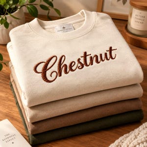 May include: A stack of four folded crewneck sweatshirts in cream, light beige, medium brown, and dark olive green. The top cream sweatshirt features the word Chestnut embroidered in dark brown script across the chest and a small interior label.