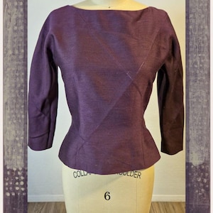 Peut inclure: Un haut à manches longues violet foncé avec une encolure bateau et une couture diagonale sur le devant. Le vêtement présente une taille ajustée et des manches trois-quarts, présenté sur un mannequin de taille 6 avec des inscriptions.