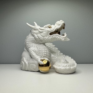 May include: A white ceramic dragon figurine featuring intricate scale detailing, a wide-open mouth with gold-colored teeth, and a glossy gold-colored sphere held firmly in one clawed hand. The dragon is depicted in a coiled, seated position.