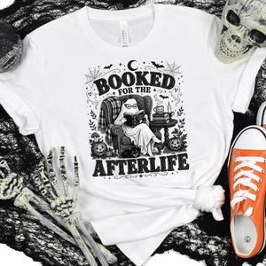 May include: A white t-shirt featuring a black graphic print of a ghost wearing glasses and reading a book in an armchair. The text reads "BOOKED FOR THE AFTERLIFE" and includes illustrations of pumpkins, bats, spiderwebs, and a stack of books.