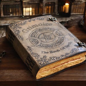 May include: A tan leather-bound book titled Grimoire The Book Of Spells with an embossed pentagram and crescent moon design on the cover. The book features metal latches, decorative floral borders, and thick, textured, cream-colored pages.