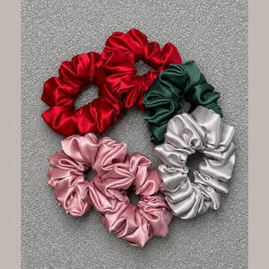 May include: A set of four satin hair scrunchies in solid colors. The collection includes one bright red scrunchie, one dark forest green scrunchie, one light dusty pink scrunchie, and one metallic silver-grey scrunchie, all featuring a gathered fabric design.