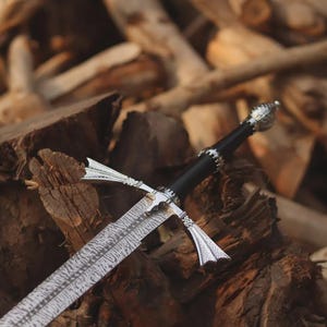 Può includere: Una spada con lama in acciaio Damasco a motivi, impugnatura nera e guardia e pomolo color argento. La guardia presenta bracci svasati a forma di ala con dettagli intricati, mentre il pomolo ha una forma arrotondata e testurizzata.