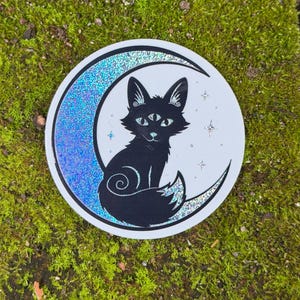 May include: A circular sticker featuring a black silhouette of a three-eyed fox sitting in front of a holographic crescent moon. The design includes small holographic star accents on a white background, creating a reflective and detailed graphic.