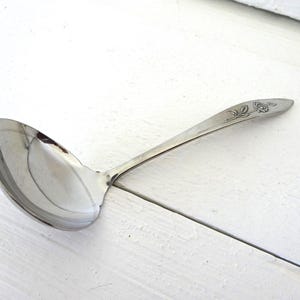 May include: A silver-toned metal soup ladle with a polished, rounded bowl and a slender handle. The handle features a delicate, embossed floral design near the tip, showcasing a single flower with leaves in a classic, understated style.