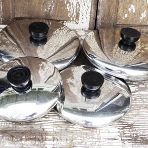 May include: Four silver-colored pot lids with black plastic handles. The lids are stacked on top of each other.