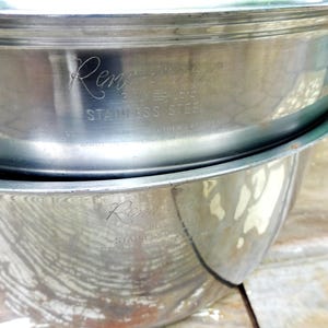 May include: Two stacked silver-colored stainless steel mixing bowls with a polished finish. The top bowl features an engraved logo that reads "Rene-Ware" followed by "STAINLESS STEEL" in capital letters. The bowls have a rounded, deep design.