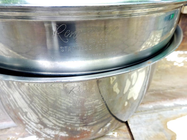 May include: Two stacked silver-colored stainless steel mixing bowls with a polished finish. The top bowl features an engraved logo that reads "Rene-Ware" followed by "STAINLESS STEEL" in capital letters. The bowls have a rounded, deep design.