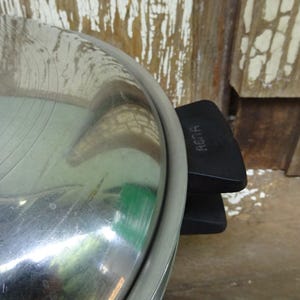 May include: A stainless steel pot lid with a polished reflective surface and a black plastic handle featuring the embossed text RENA. The lid has a circular design with a metallic rim and a sturdy grip attached to the side for easy handling.