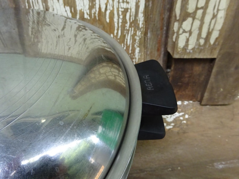 May include: A stainless steel pot lid with a polished reflective surface and a black plastic handle featuring the embossed text RENA. The lid has a circular design with a metallic rim and a sturdy grip attached to the side for easy handling.