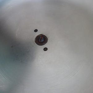 May include: A circular, brushed silver-toned metal surface featuring a central dark, oxidized circular mounting point with a single screw head. Two small, perfectly round holes are positioned symmetrically on either side of the central hardware.