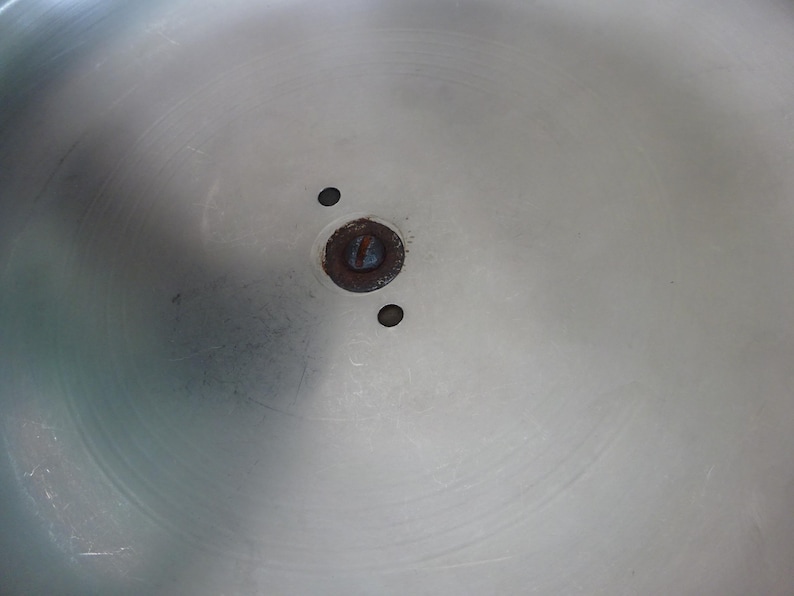 May include: A circular, brushed silver-toned metal surface featuring a central dark, oxidized circular mounting point with a single screw head. Two small, perfectly round holes are positioned symmetrically on either side of the central hardware.